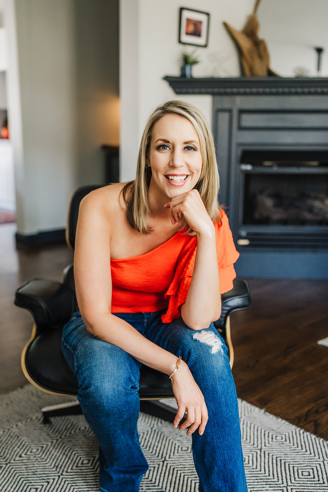 Nicki Krawczyk, founder of Nicki K Media, sits in a black swivel chair in jeans and a one-shoulder orange top